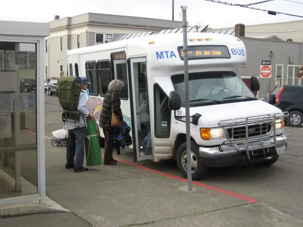 A small bus labeled "MTA" is at a stop, with passengers boarding, including one with a large backpack and another with bags.