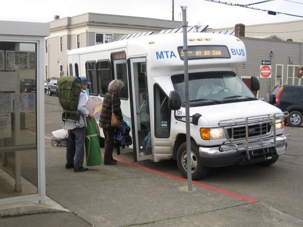 A small bus labeled "MTA" is at a stop, with passengers boarding, including one with a large backpack and another with bags.