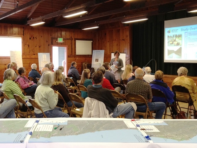 A community meeting in a wooden hall, with attendees listening to a speaker and presentations displayed on a screen.