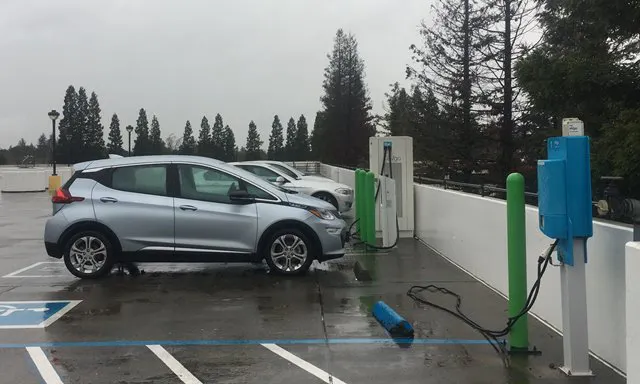 Two electric cars are parked at a charging station on a rainy day, with charging equipment visible nearby.