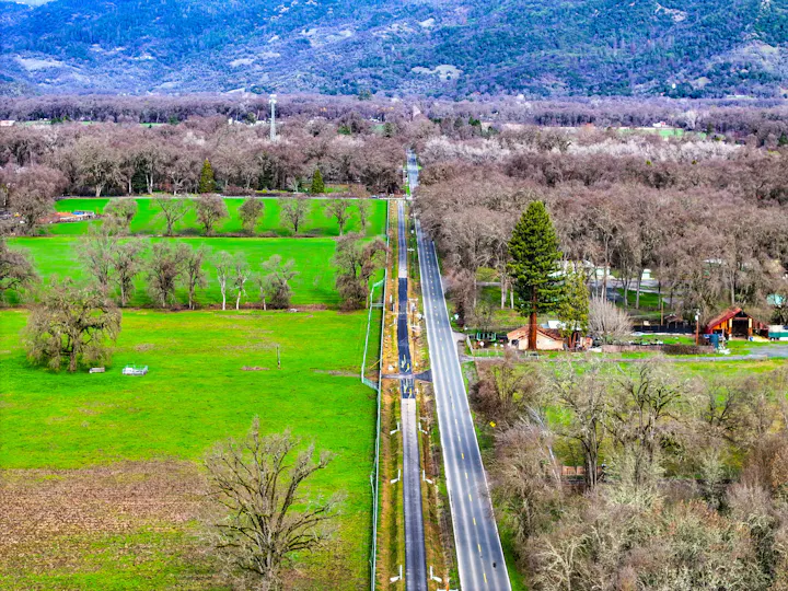 Aerial view of a straight road with trail construction along one side through green fields, flanked by trees and hills in the background.