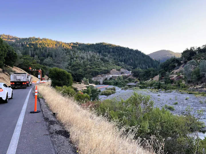 A scenic highway view with trees, a river, traffic cones, and vehicles, set against a mountainous backdrop at dusk.