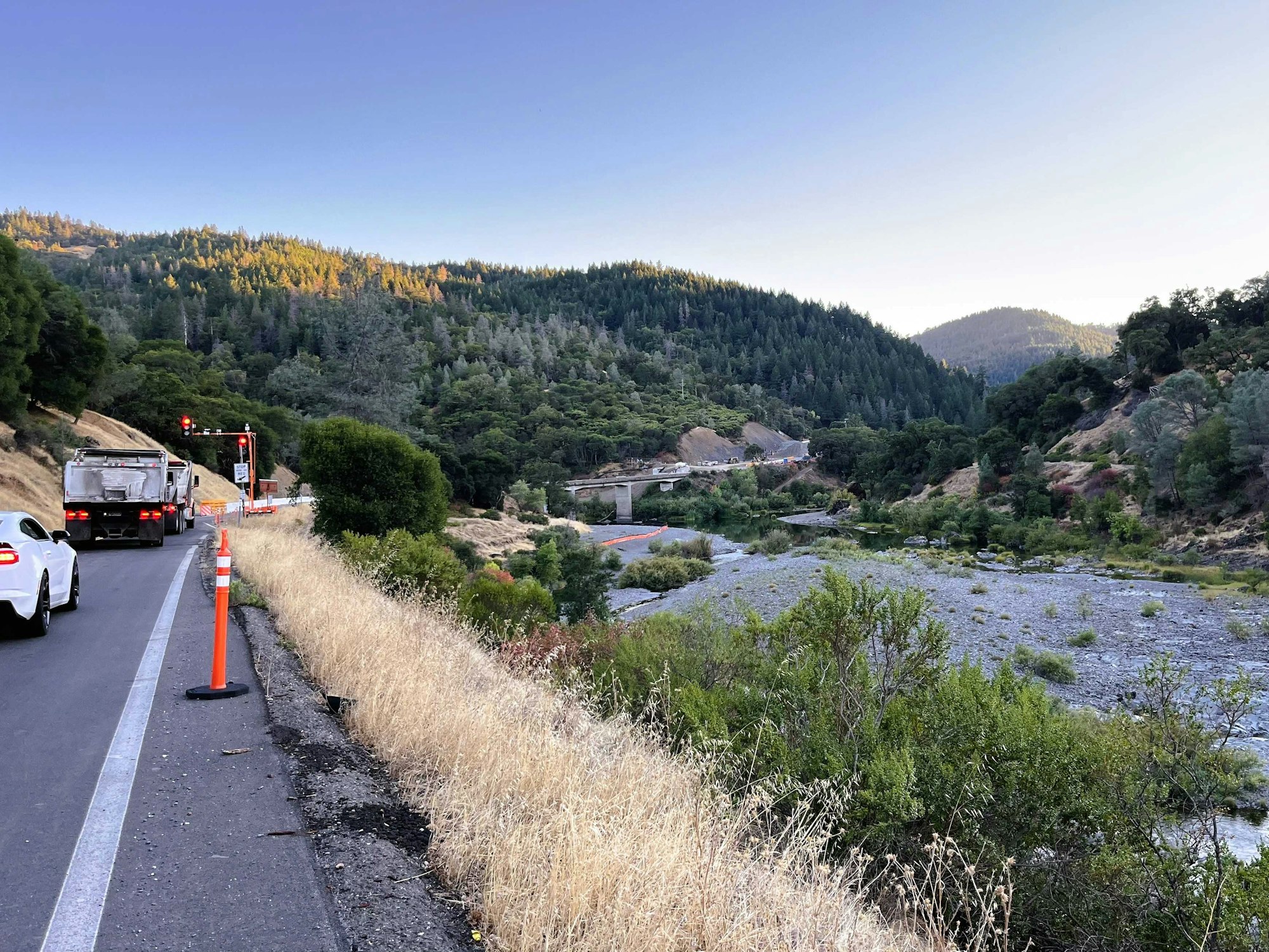 A scenic highway view with trees, a river, traffic cones, and vehicles, set against a mountainous backdrop at dusk.