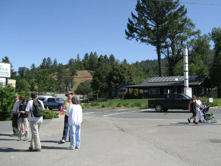 A sunny day scene showing a group of people conversing near a motel, with a truck parked and trees in the background.
