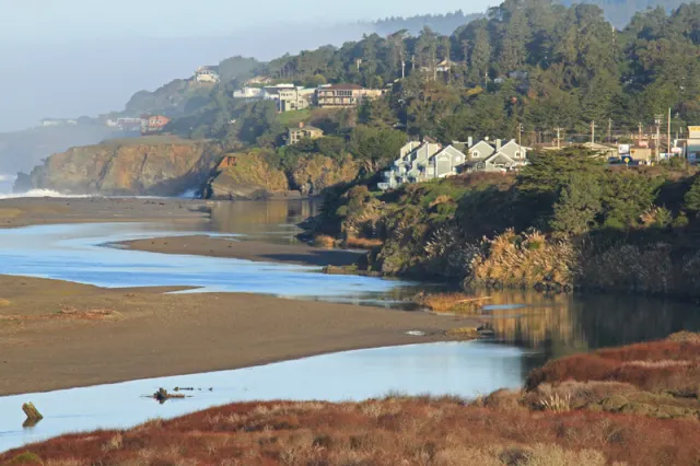A serene coastal scene featuring a river, greenery, and houses along the hillside, with gentle waves in the background.