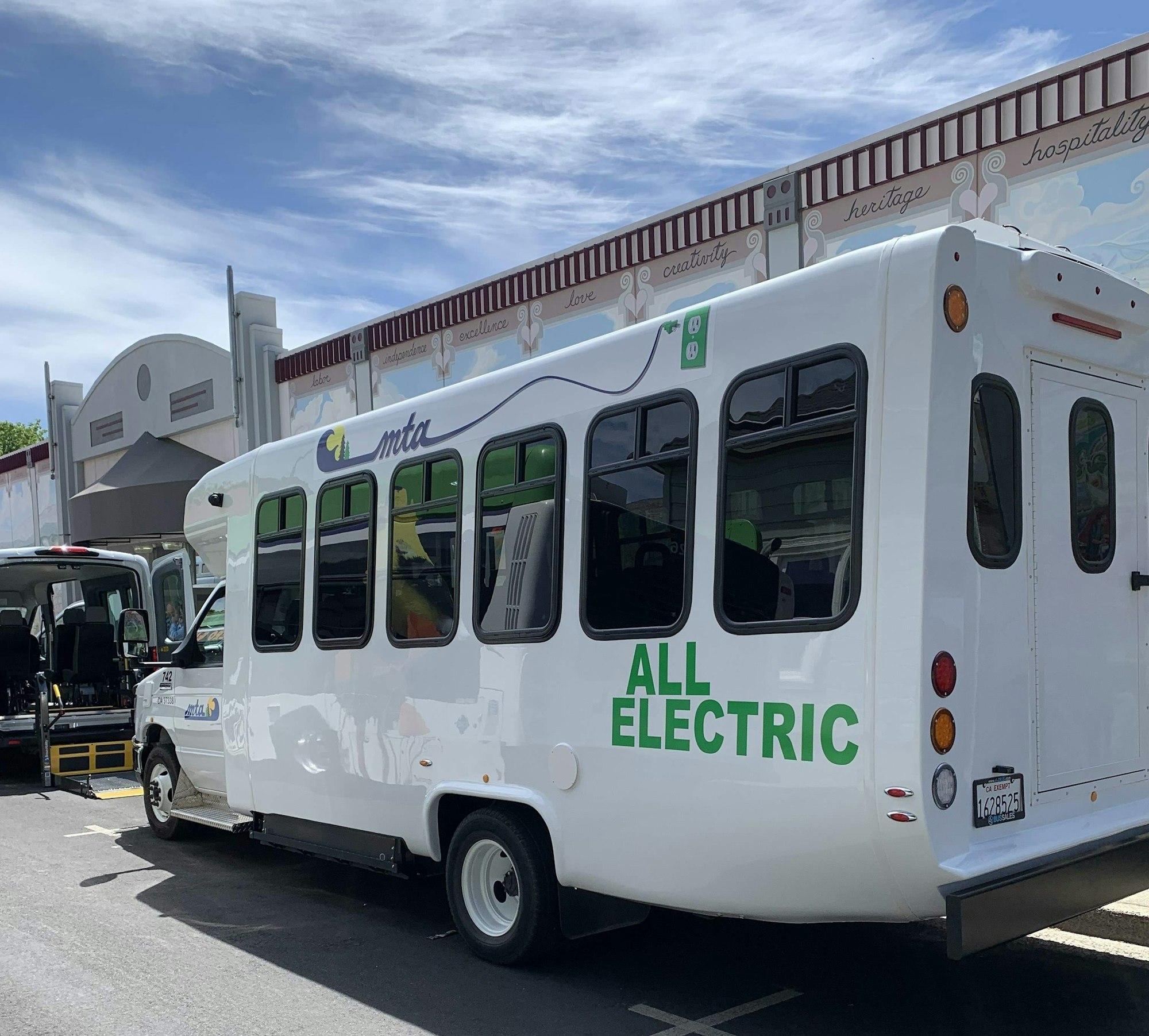 All-electric bus parked in front of a building with mural and blue sky.
