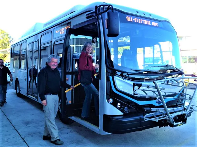 An all-electric bus is shown, with a woman boarding and a man smiling nearby, highlighting eco-friendly transportation.