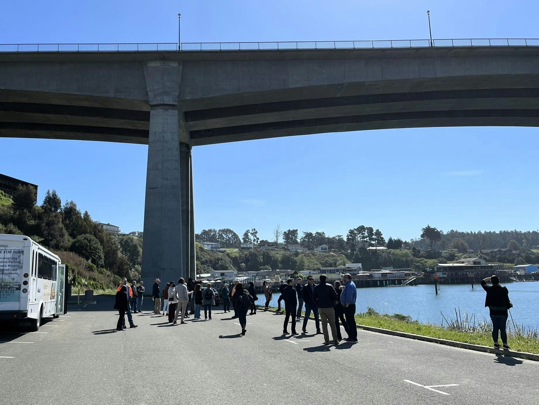 A group of people standing near a bus under a large concrete bridge by the water on a sunny day.