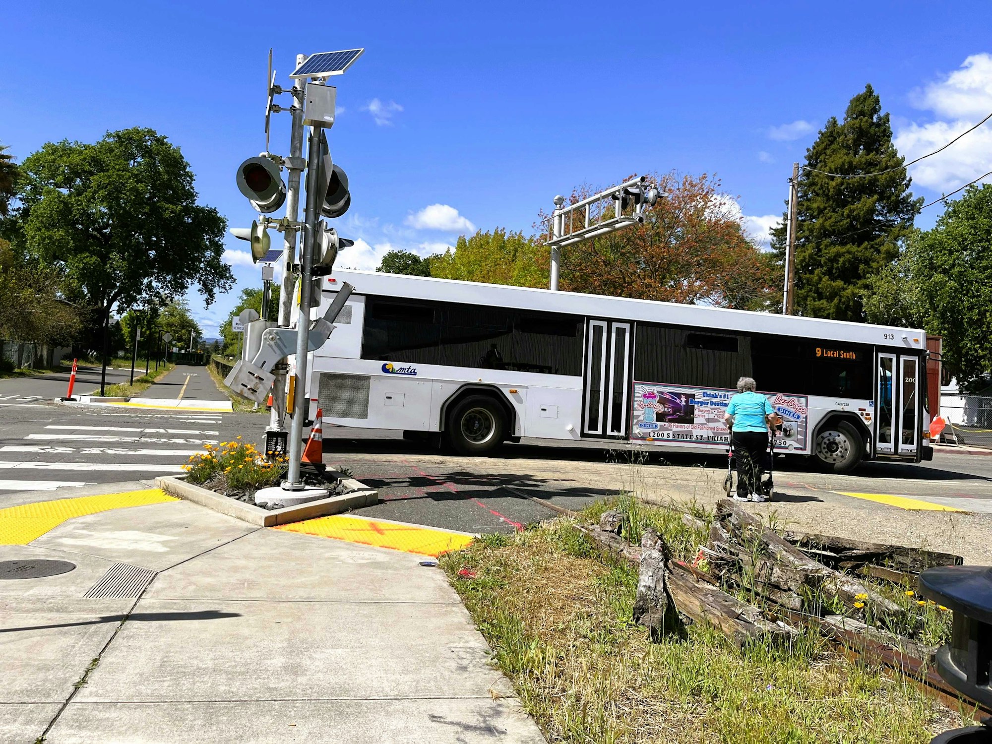 A city bus approaches a train crossing, with a woman walking nearby. Clear blue sky, greenery, and traffic signals are visible.