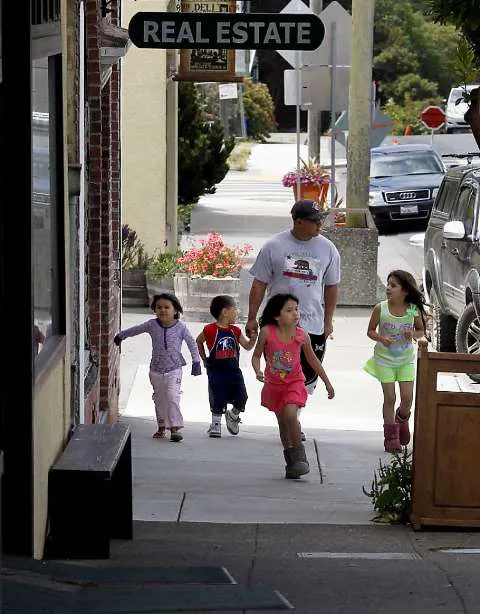 A man walks with four children down a sidewalk, passing a "REAL ESTATE" sign and parked cars.
