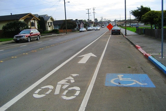 A street featuring a bike lane with a bike symbol, a wheelchair accessibility symbol, and a construction warning sign.