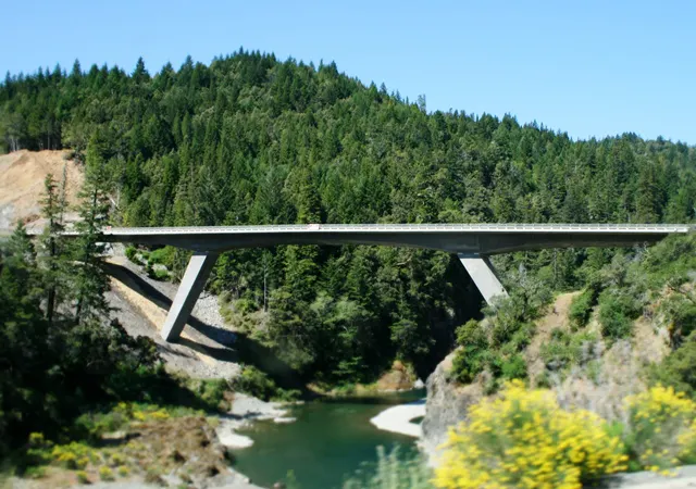 A concrete bridge spans over a river, surrounded by dense greenery and forested hills under a clear blue sky.