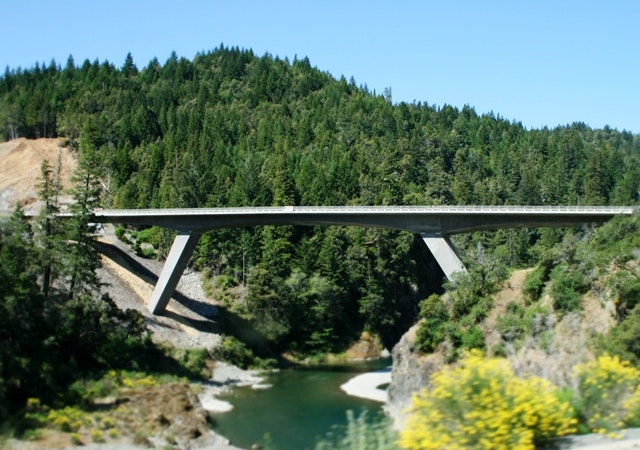A concrete bridge spans over a river, surrounded by dense greenery and forested hills under a clear blue sky.