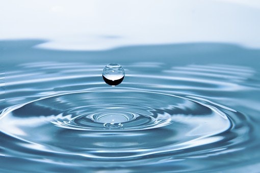 A single water droplet creates ripples on a calm water surface.