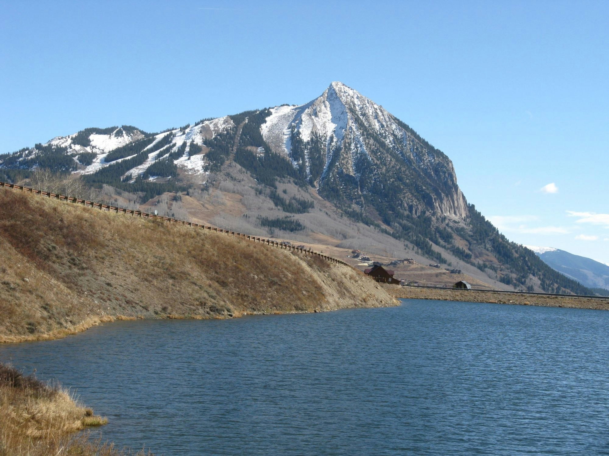 A mountain with snow patches overlooking a tranquil lake and a clear blue sky.