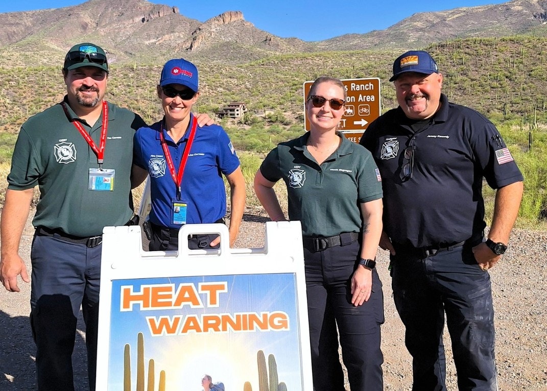 Four people in uniform pose by a "Heat Warning" sign in a desert area with mountains in the background.