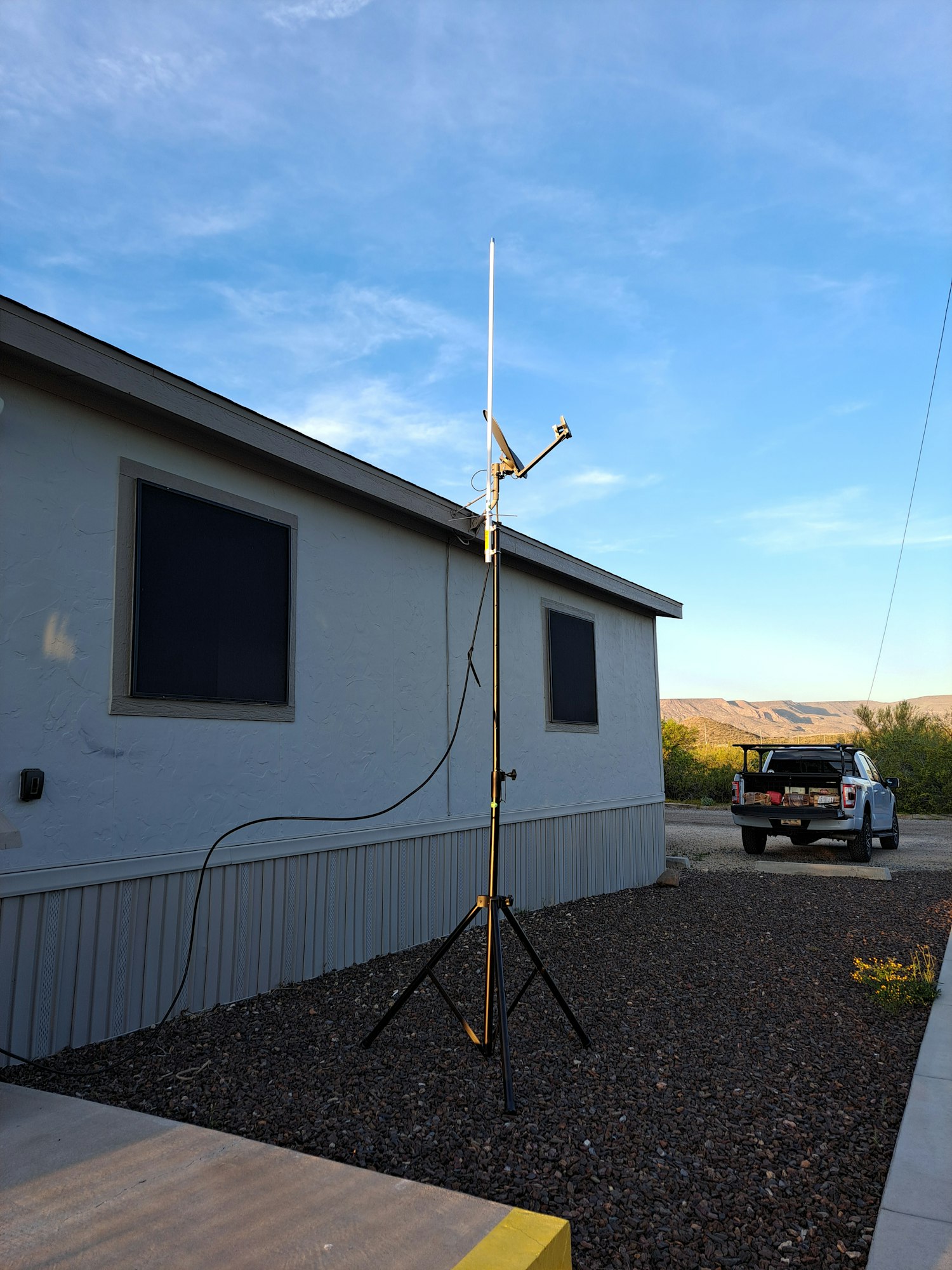A tall antenna on a tripod stand near a building, with a parked vehicle in the background under a clear blue sky.