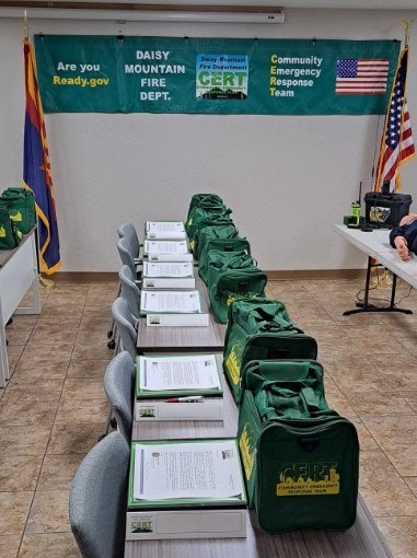 Tables with green bags and folders set up for a Community Emergency Response Team meeting by the Daisy Mountain Fire Dept. Flags are displayed.