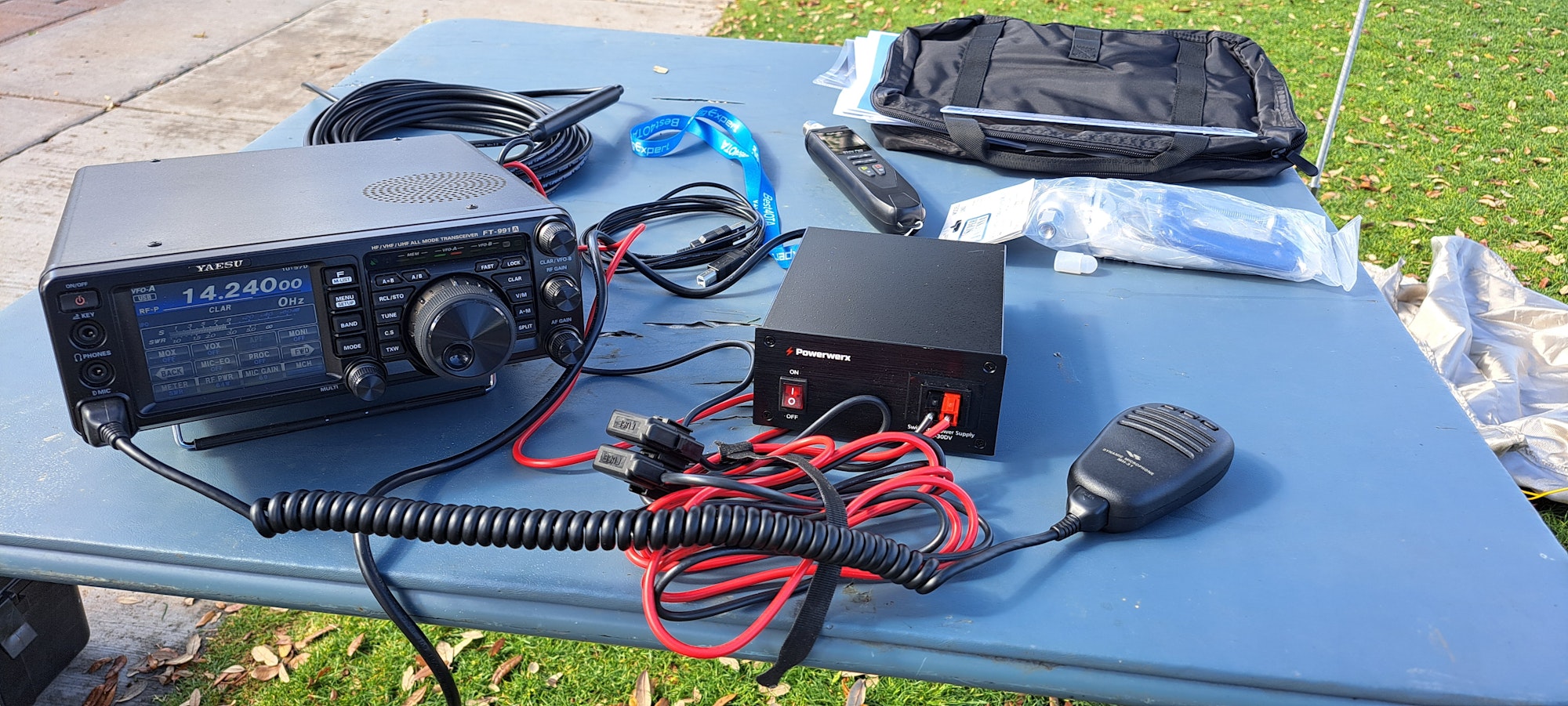 Ham radio setup with transceiver, microphone, cables, and power supply on a table outdoors.