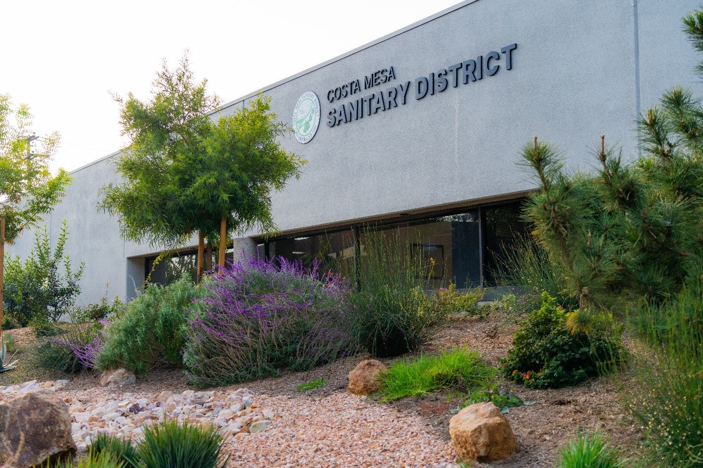 Building facade labeled "Costa Mesa Sanitary District" with landscaping, trees, and shrubs in front.