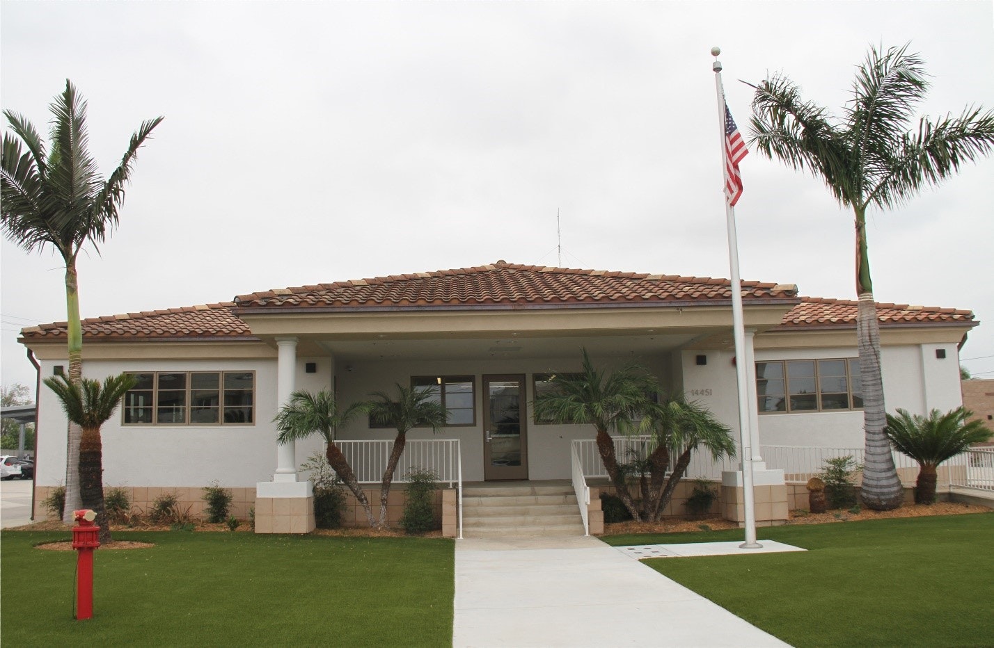 A single-story building with a tiled roof, surrounded by palm trees and grass, featuring a flagpole with the American flag.