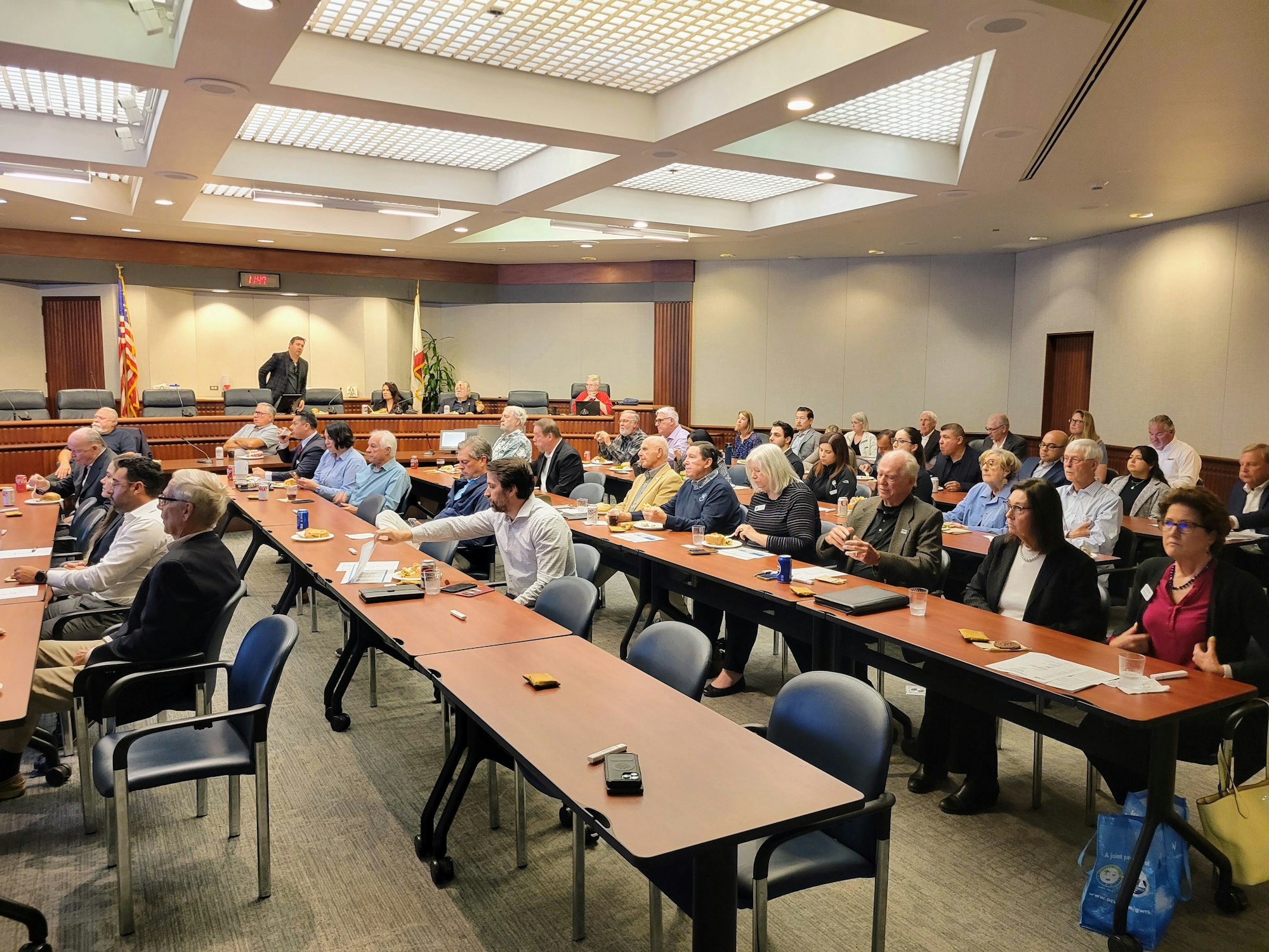 A conference room with people attending a meeting, seated at long tables with papers and drinks, under bright ceiling lights.