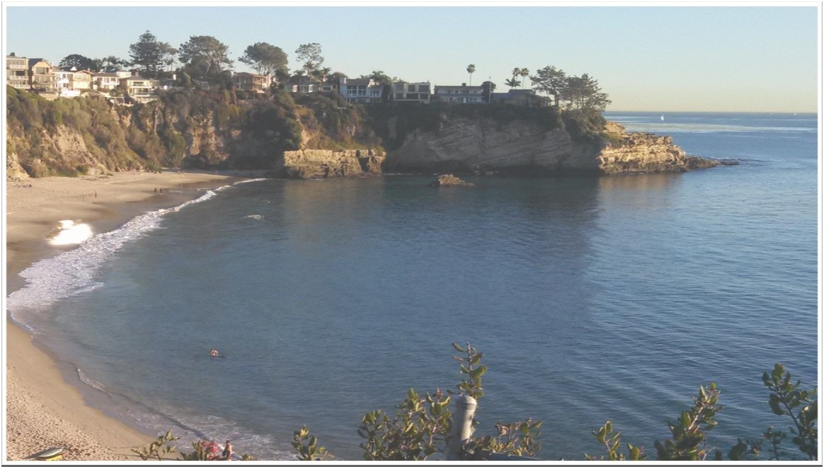 A beach with gentle waves, a cliff with houses, and calm blue waters with a promontory.