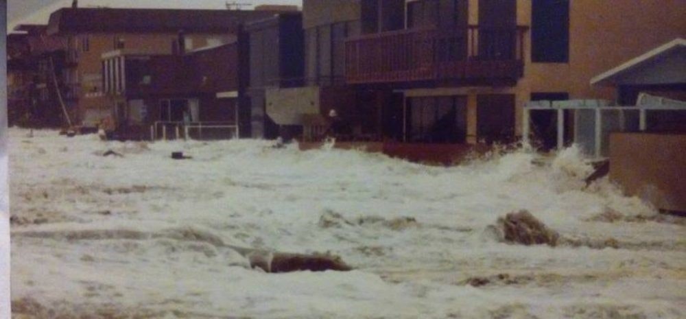 Flooded street with water surging through, impacting buildings.
