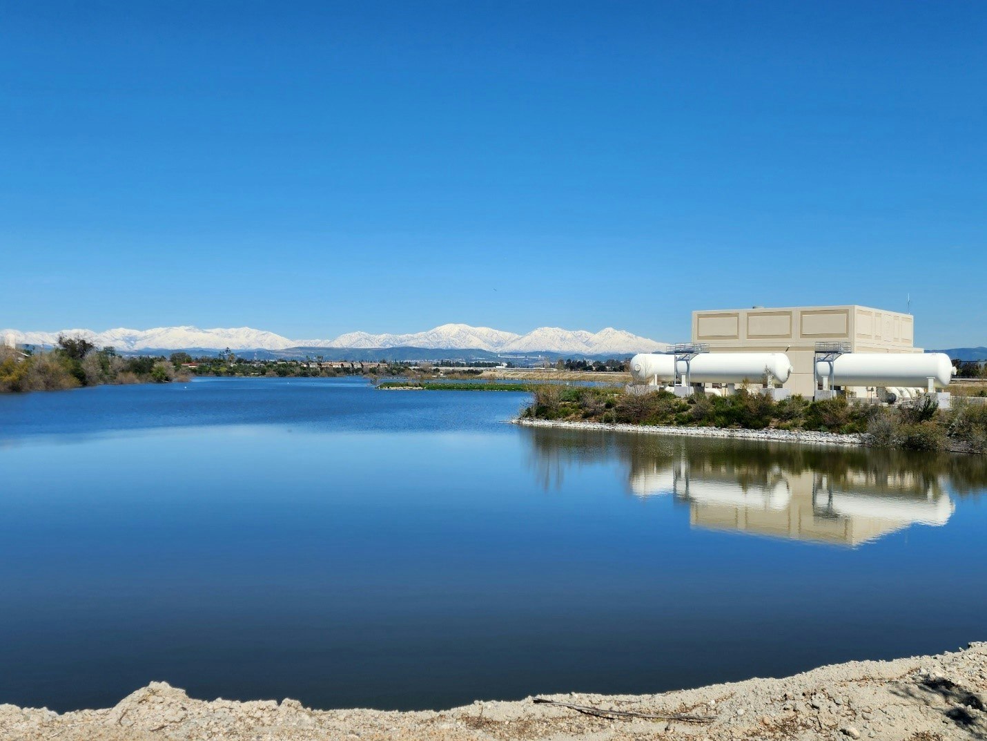 A building with cylindrical tanks beside a calm lake, snowy mountains in the background, clear blue sky.