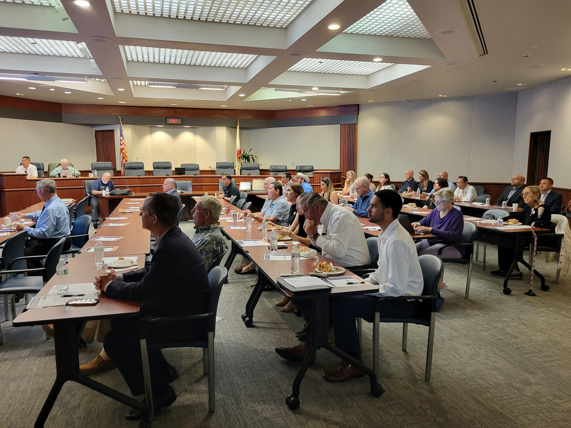 A group of people in a meeting room with papers and food, listening to a speaker.