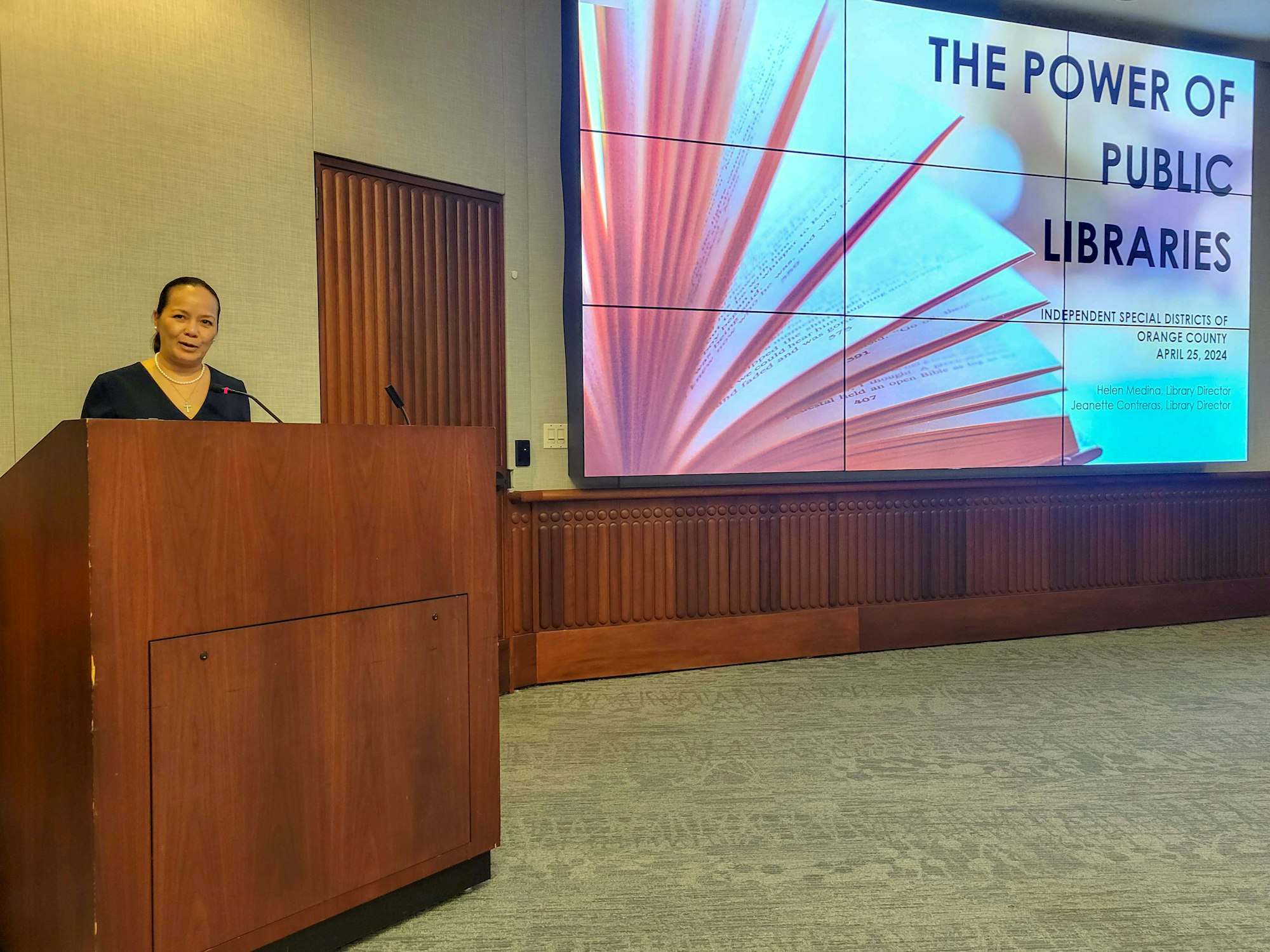 A person stands at a lectern in a conference room with a presentation titled "THE POWER OF PUBLIC LIBRARIES" in the background.