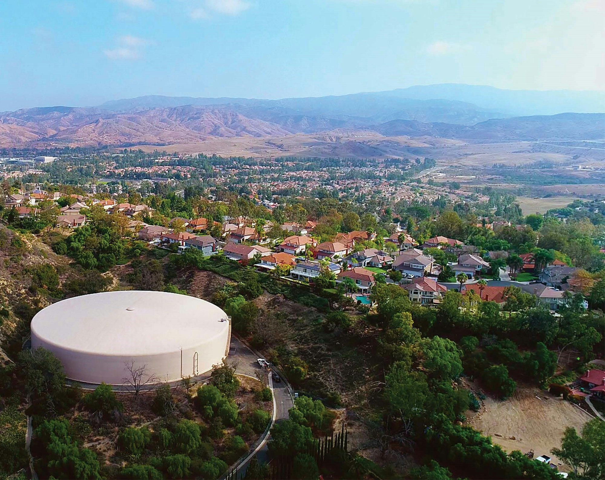 Aerial view of a suburban area with houses, a large water tank, greenery, and mountains in the background.