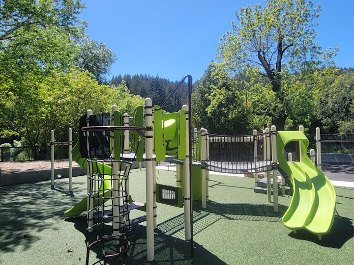 A playground with slides and climbing structures on a sunny day, surrounded by trees.