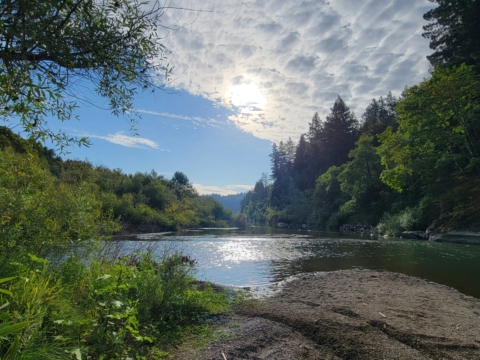 A serene river scene surrounded by lush greenery and trees under a partly cloudy sky, reflecting sunlight on the water.
