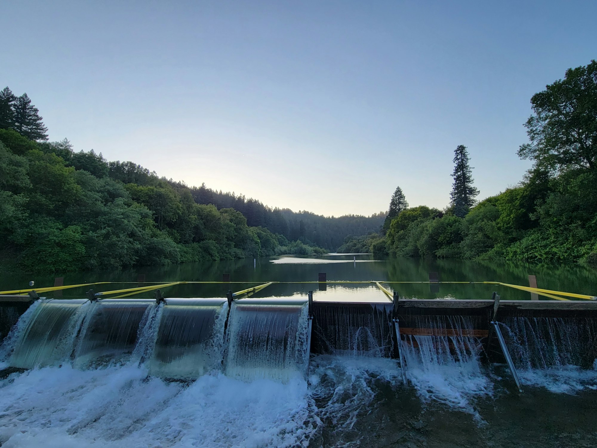 A serene lake with trees and a small dam with cascading water at twilight.