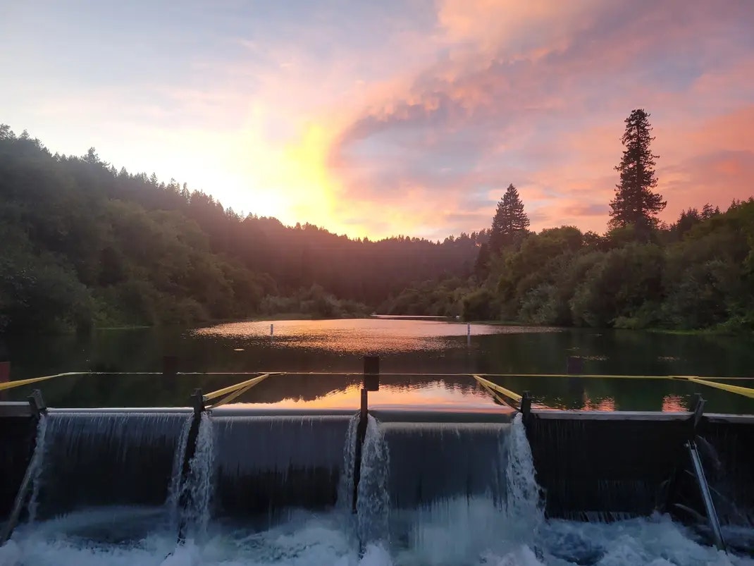 A serene sunset over a forested area, with water cascading down a small dam into a calm river.