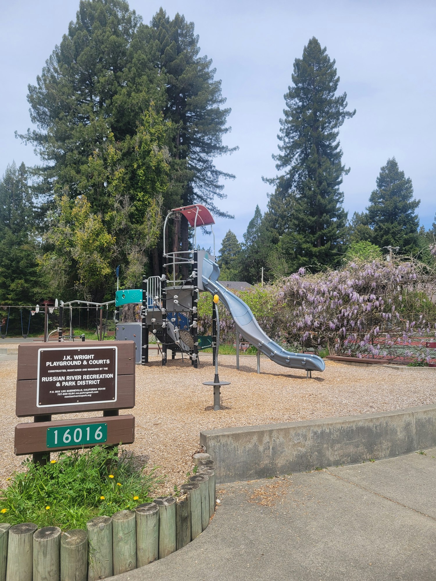 A playground with slide, swing, and signage, flanked by trees and a clear sky. (Note: Image is rotated.)