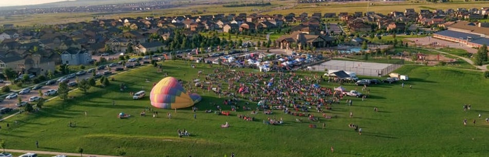 Aerial view of a large gathering in a park with a colorful hot air balloon on the ground, surrounded by houses and open space.