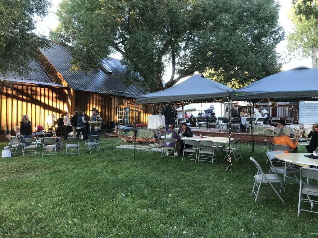 Outdoor gathering with people sitting and standing, tables under canopies, near a wooden building, surrounded by trees.
