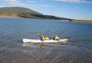 Two people kayaking on a calm lake with hills in the background under a blue sky.