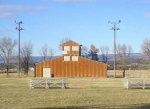 A rustic barn with a central tower, surrounded by open fields and trees, under a clear blue sky.