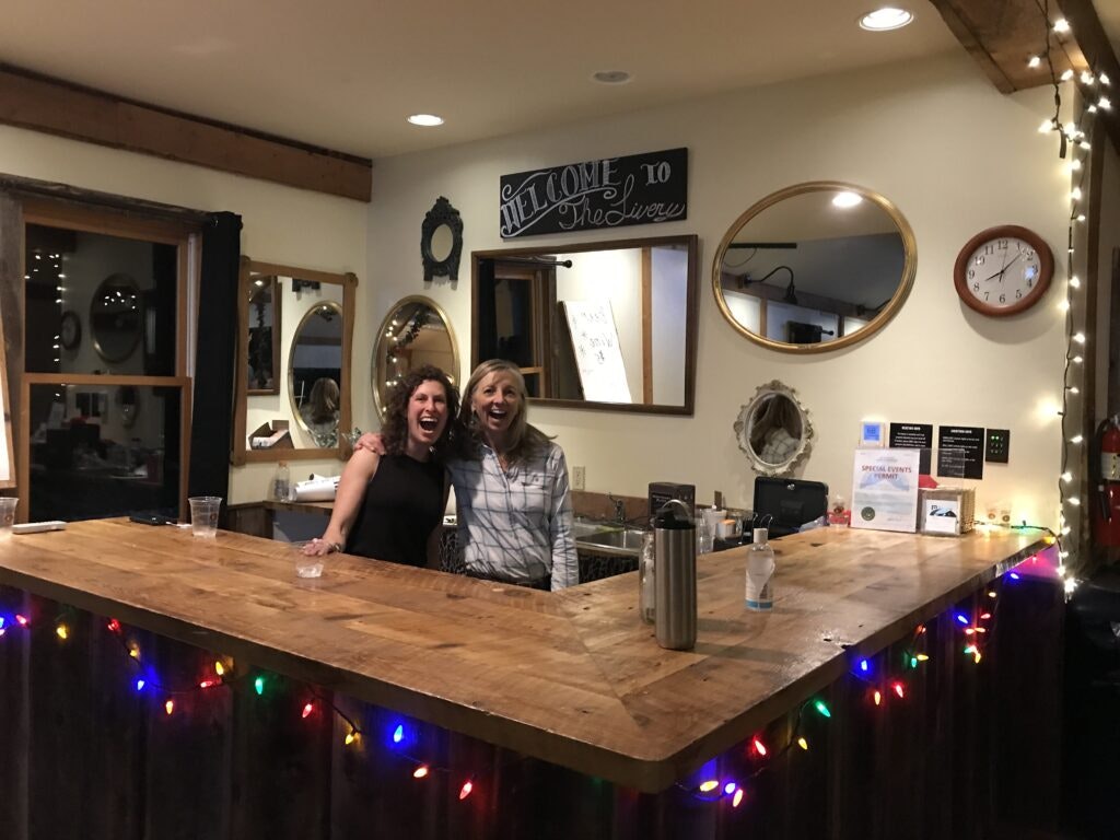 Two people smiling behind a bar decorated with colorful lights, with mirrors and a "Welcome" sign on the wall.