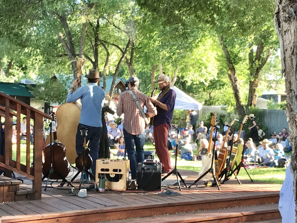 A band performs on an outdoor stage under trees, with guitars and other instruments. Audience seated on the grass in the background.