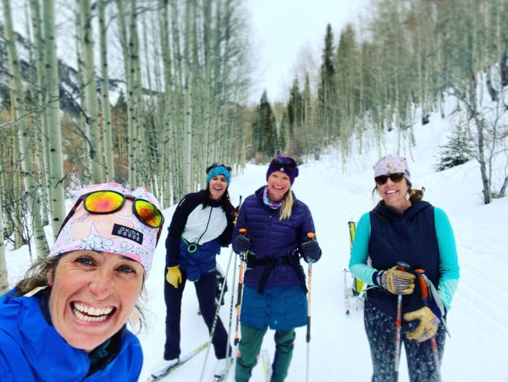 Four women smiling while skiing on a snowy trail lined with trees.