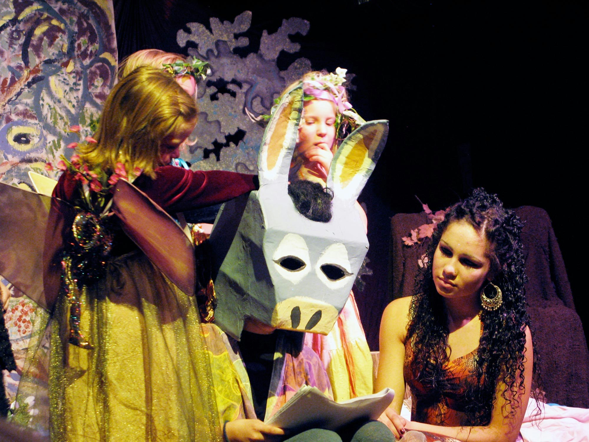 Children in costumes performing a play, with a person wearing a donkey mask, surrounded by colorful scenery and props.