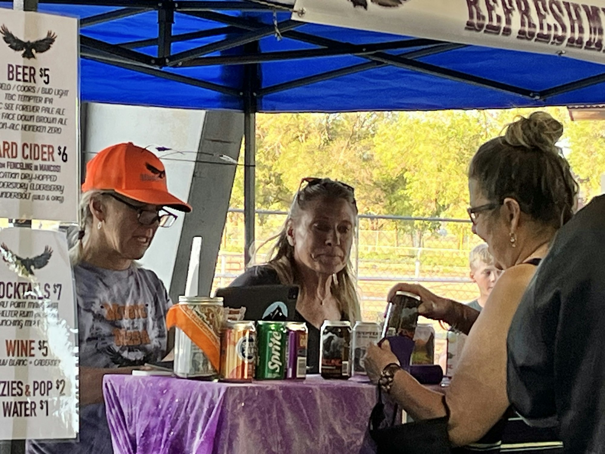 People at a refreshment stand under a blue canopy, with drinks and a menu listing beer, cider, cocktails, wine, and soda.