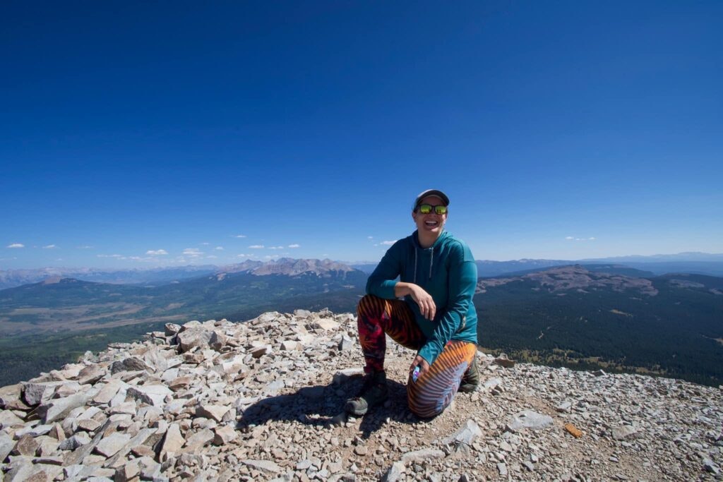 Person smiling atop a rocky mountain peak with a vast landscape and clear blue sky in the background.