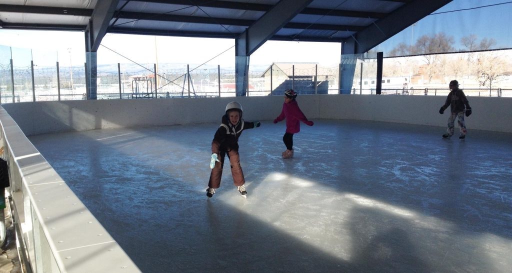 Children ice skating in an indoor rink with helmets and winter clothing.
