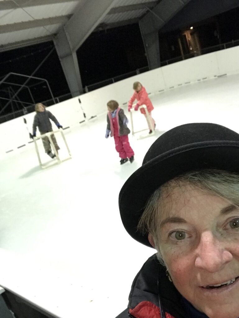 A person taking a selfie at an ice rink with three children using skating aids in the background.