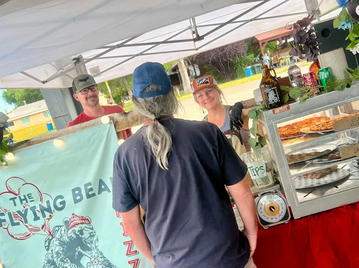 A food stall with smiling staff serving pizza to a customer, featuring a colorful banner and a variety of food options.